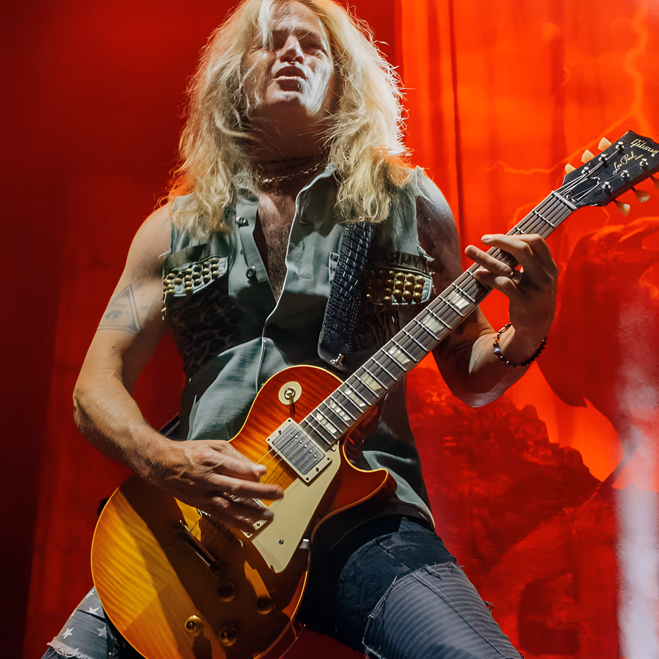 Rock musician with long blond hair playing a sunburst electric guitar under red stage lighting.