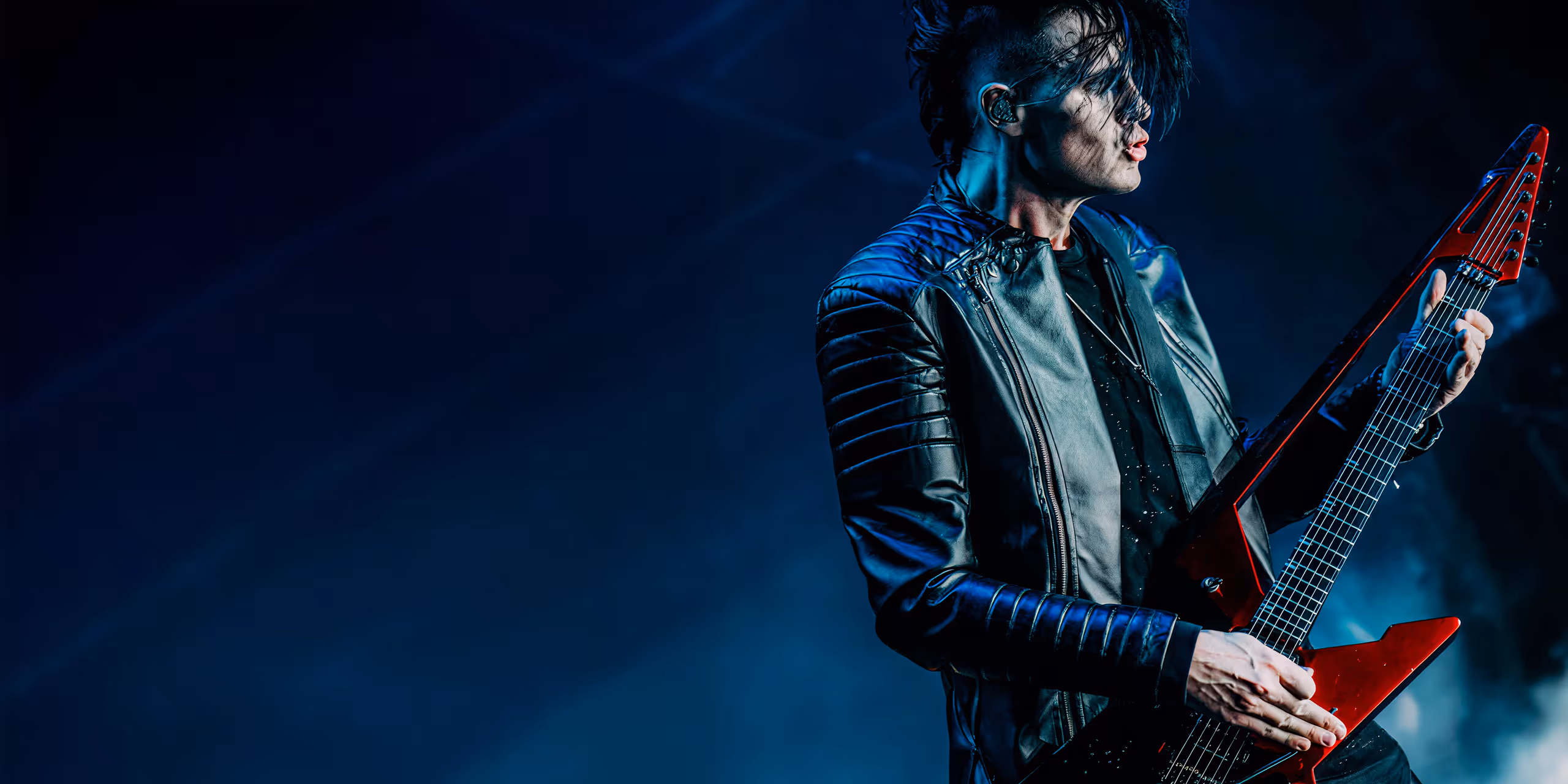 Musician with black hair playing a red electric guitar on stage under blue lighting.