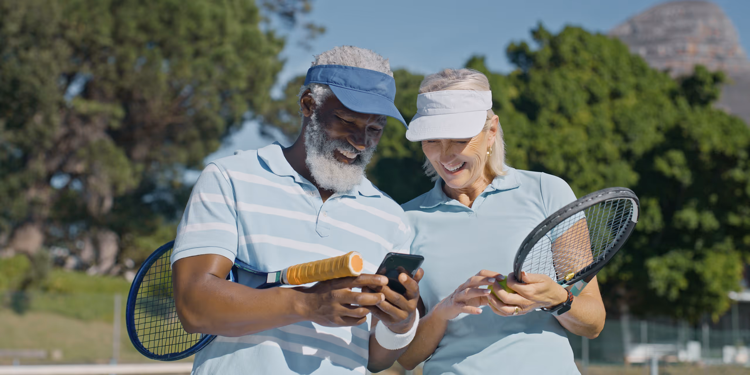 Smiling elderly man and woman in tennis attire looking at a phone together on an outdoor tennis court.