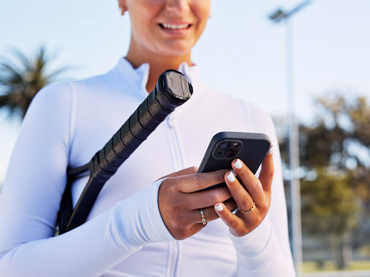 Smiling woman in white sportswear holding a tennis racquet over her shoulder while using a smartphone outdoors.