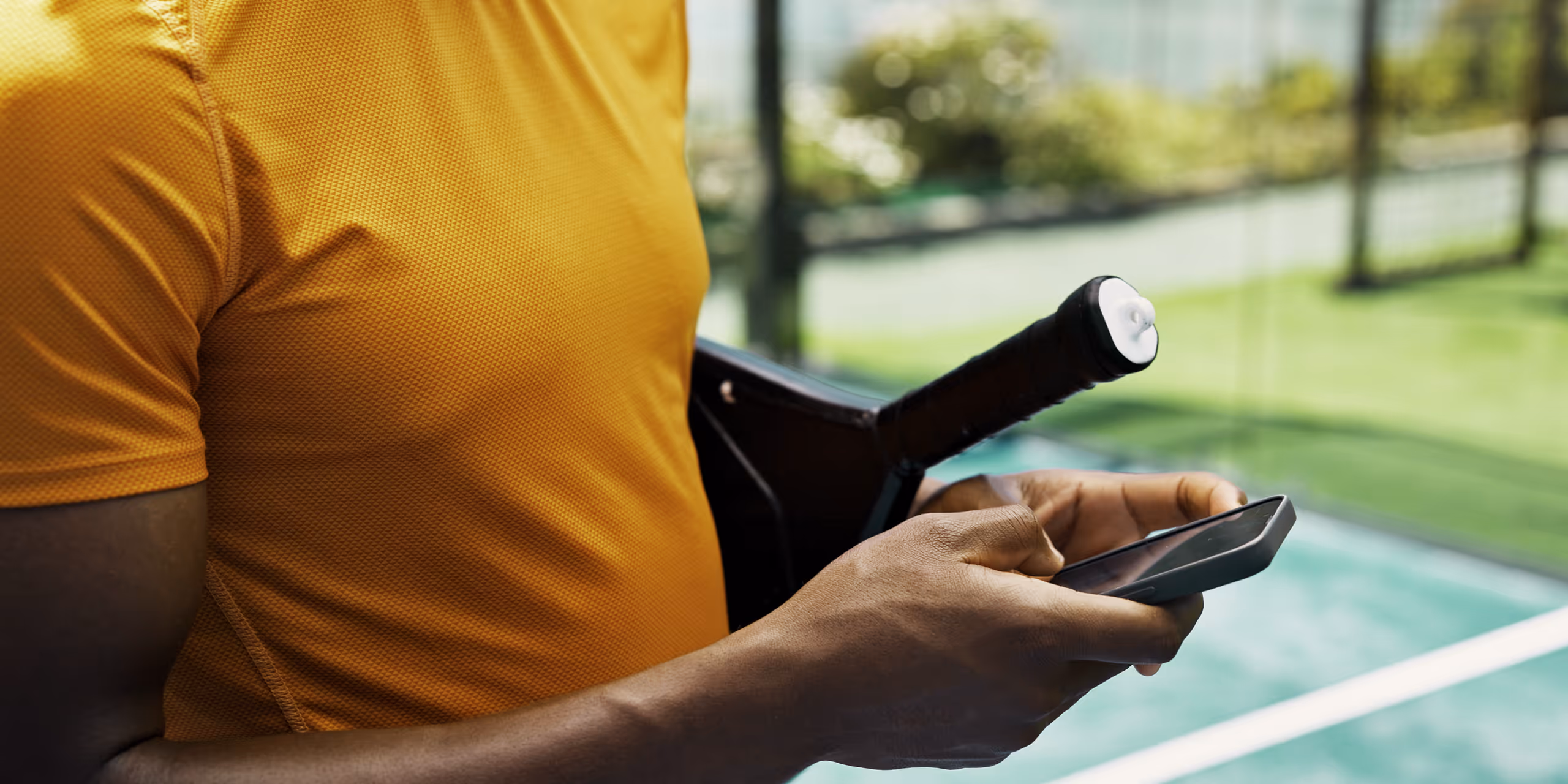 Person in an orange shirt holding a paddle and using a smartphone on a paddle tennis court.