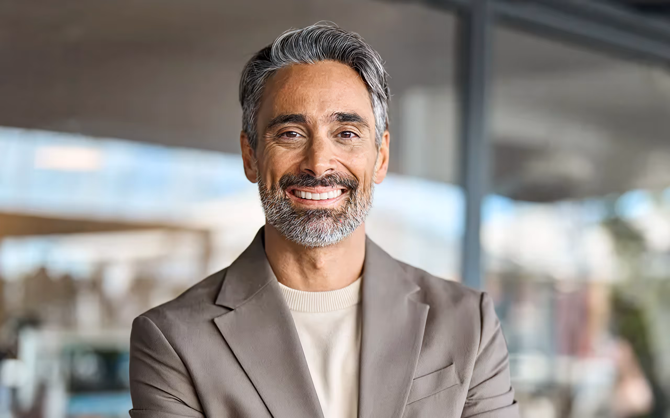 Smiling middle-aged man with gray hair and beard wearing a beige blazer and light shirt indoors.