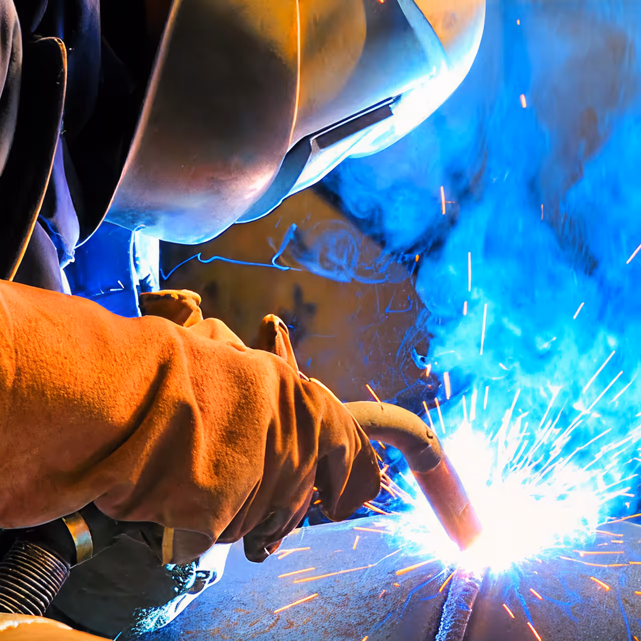 Welder wearing protective gloves and helmet welding metal pipe, bright sparks and blue smoke visible.