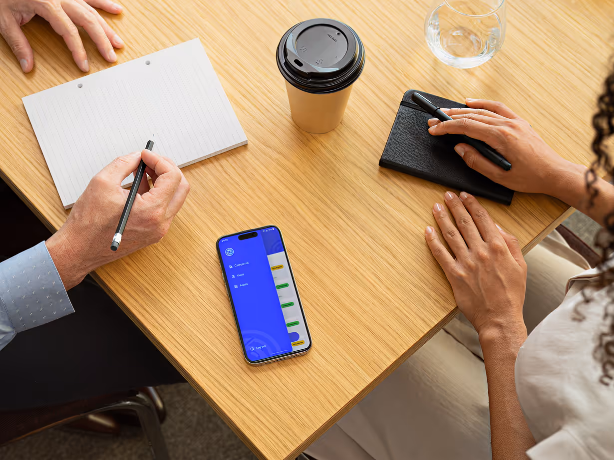 Two people at a wooden table with notebooks, pens, coffee cup, and a phone showing a blue app menu.