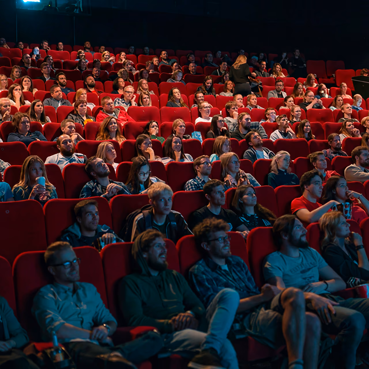 Audience of diverse adults and young adults seated in red theater seats watching a performance or movie attentively in a dimly lit auditorium.