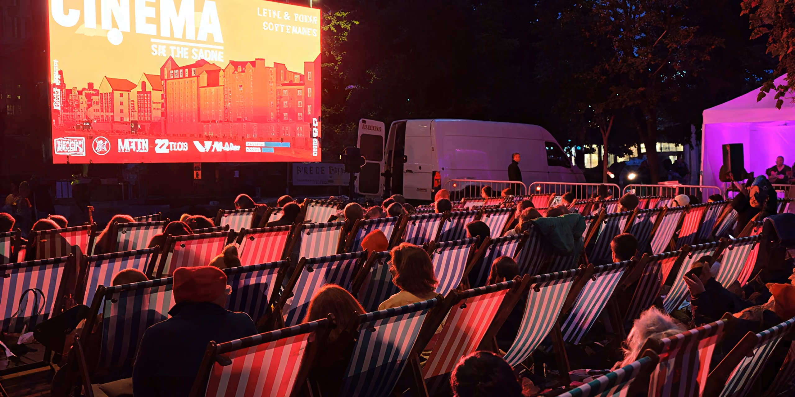 Outdoor movie screening at night with an audience seated in striped deck chairs facing a large illuminated screen showing a film titled 'Cinema'.