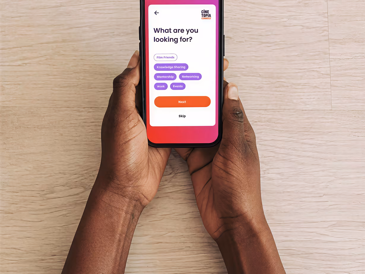 Hands holding a smartphone displaying a mobile app screen with the question 'What are you looking for?' and options including Film Friends, Knowledge Sharing, Mentorship, Networking, Work, and Events, along with a red 'Next' button and a 'Skip' link beneath.