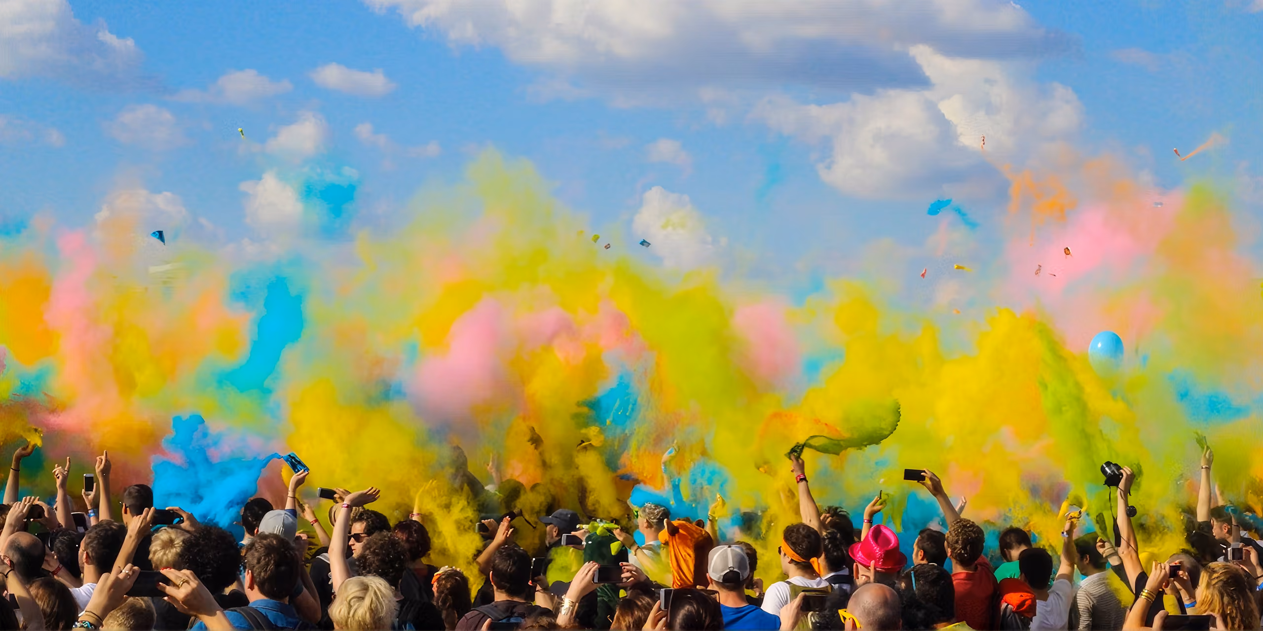 Crowd of people celebrating outdoors with vibrant clouds of colored powder in blue, yellow, pink, and green against a partly cloudy sky, many taking photos or videos with smartphones.