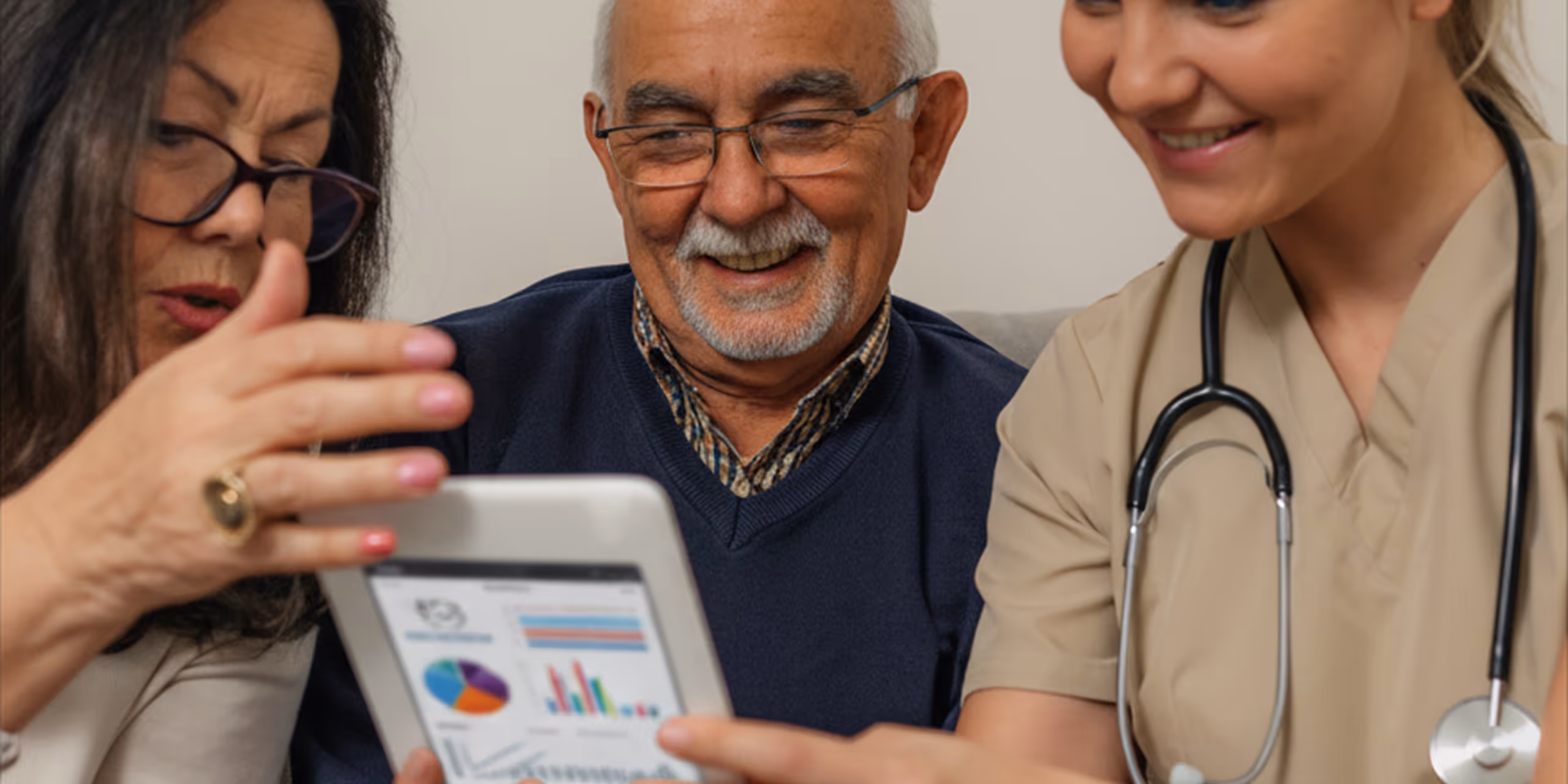 Smiling elderly man and a woman in medical scrubs with a stethoscope looking at a tablet displaying colorful charts and graphs, while another woman gestures towards the screen.