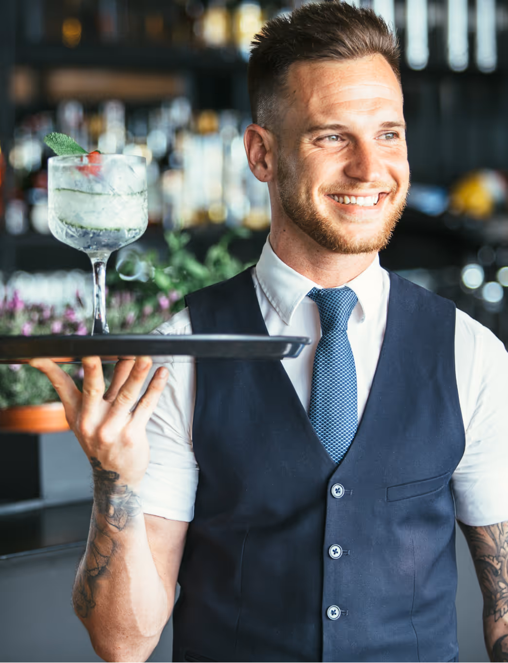 Serveur souriant en tenue professionnelle servant un cocktail dans un restaurant moderne. Image représentative des nombreuses opportunités d'emploi saisonnier dans l'hôtellerie-restauration. Les établissements recrutent intensivement des saisonniers pour la haute saison touristique : serveurs, barmen, commis, chefs de rang. Ces jobs saisonniers offrent une expérience enrichissante dans le service client et la restauration, avec possibilité d'emploi saisonnier logé dans les zones touristiques. Secteur dynamique proposant de nombreux postes de travail saisonnier pour étudiants et professionnels.