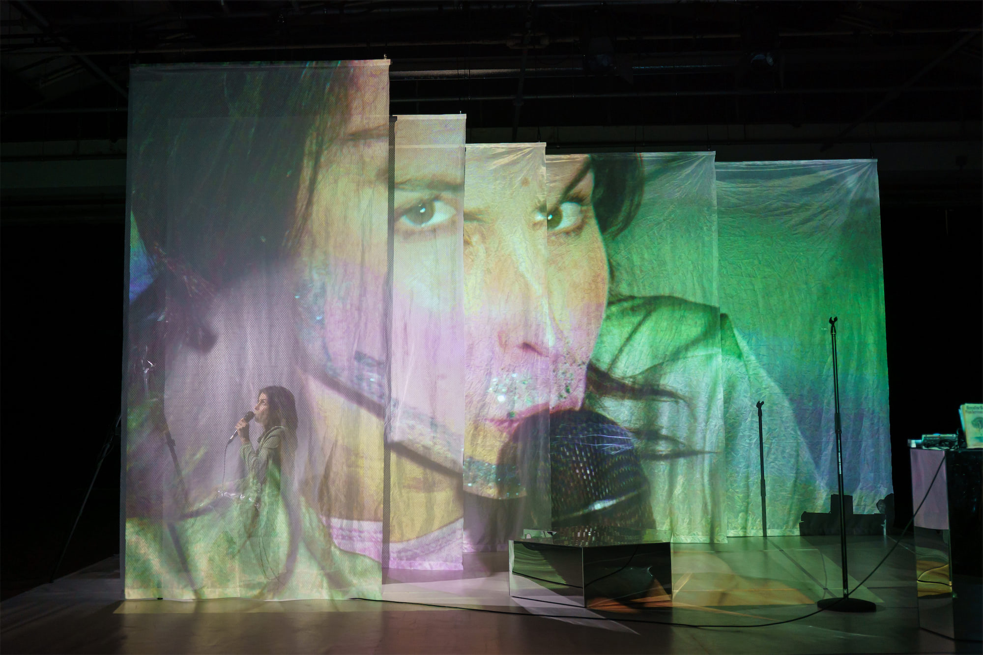 een beeld van een een persoon linksvoor die in een camera kijkt , achterop transparante doeken waarop dit beeld wordt geprojecteerd.