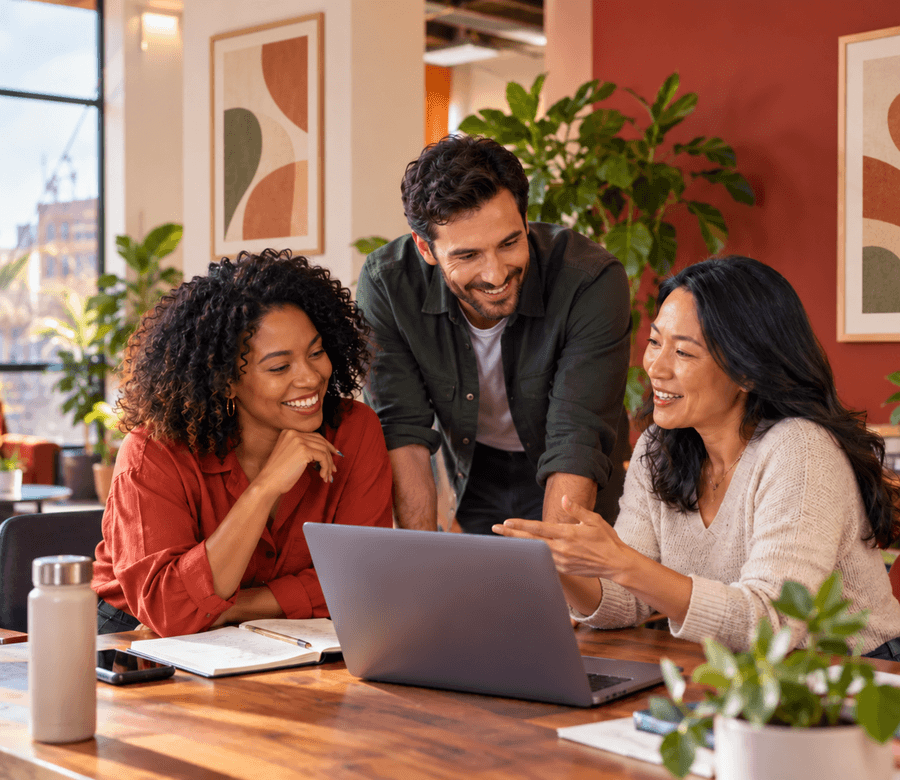 Three colleagues smiling around a laptop at a wooden table during an ongoing coaching session