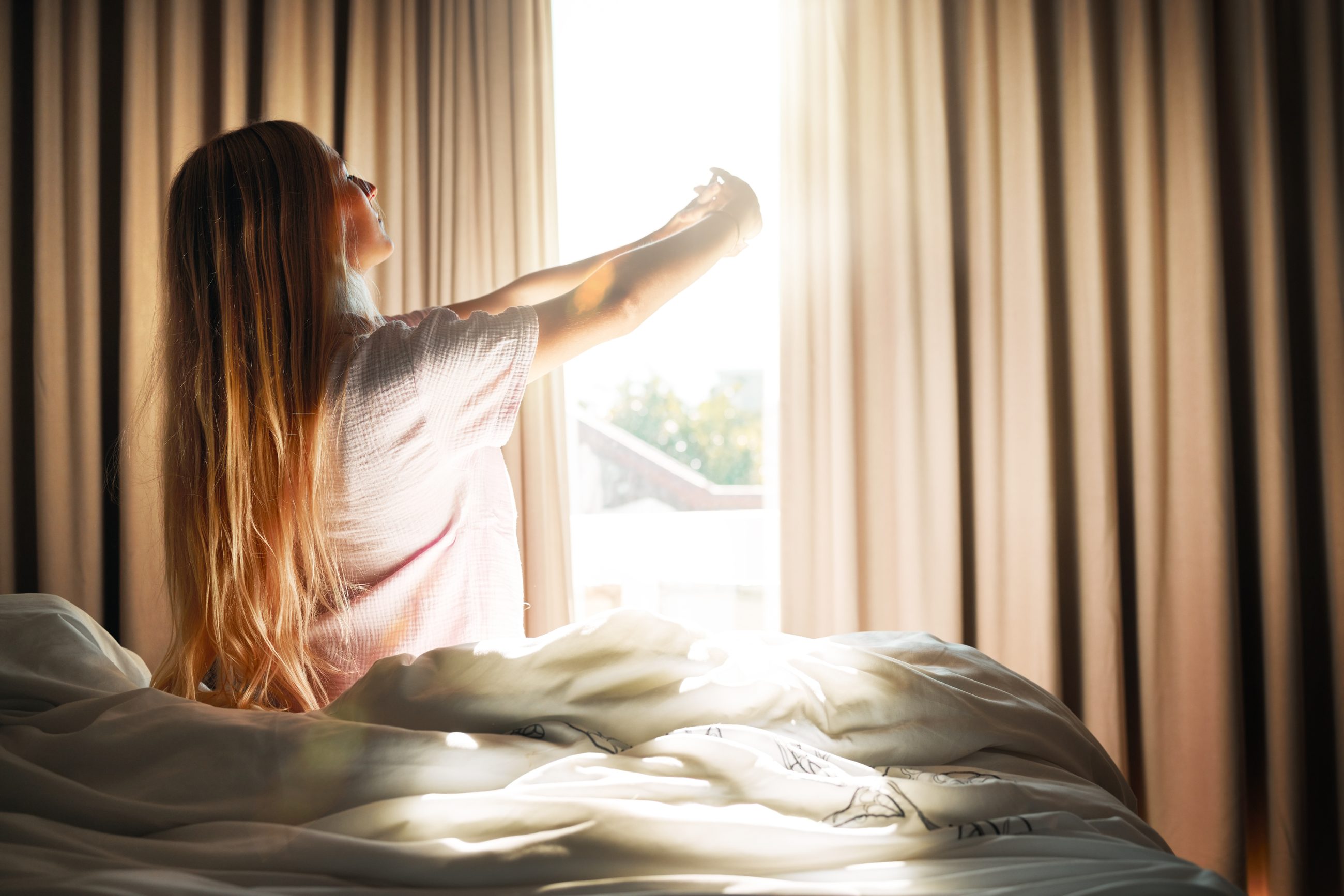Frau steht am Fenster im Schlafzimmer, Sonnenlicht fällt herein