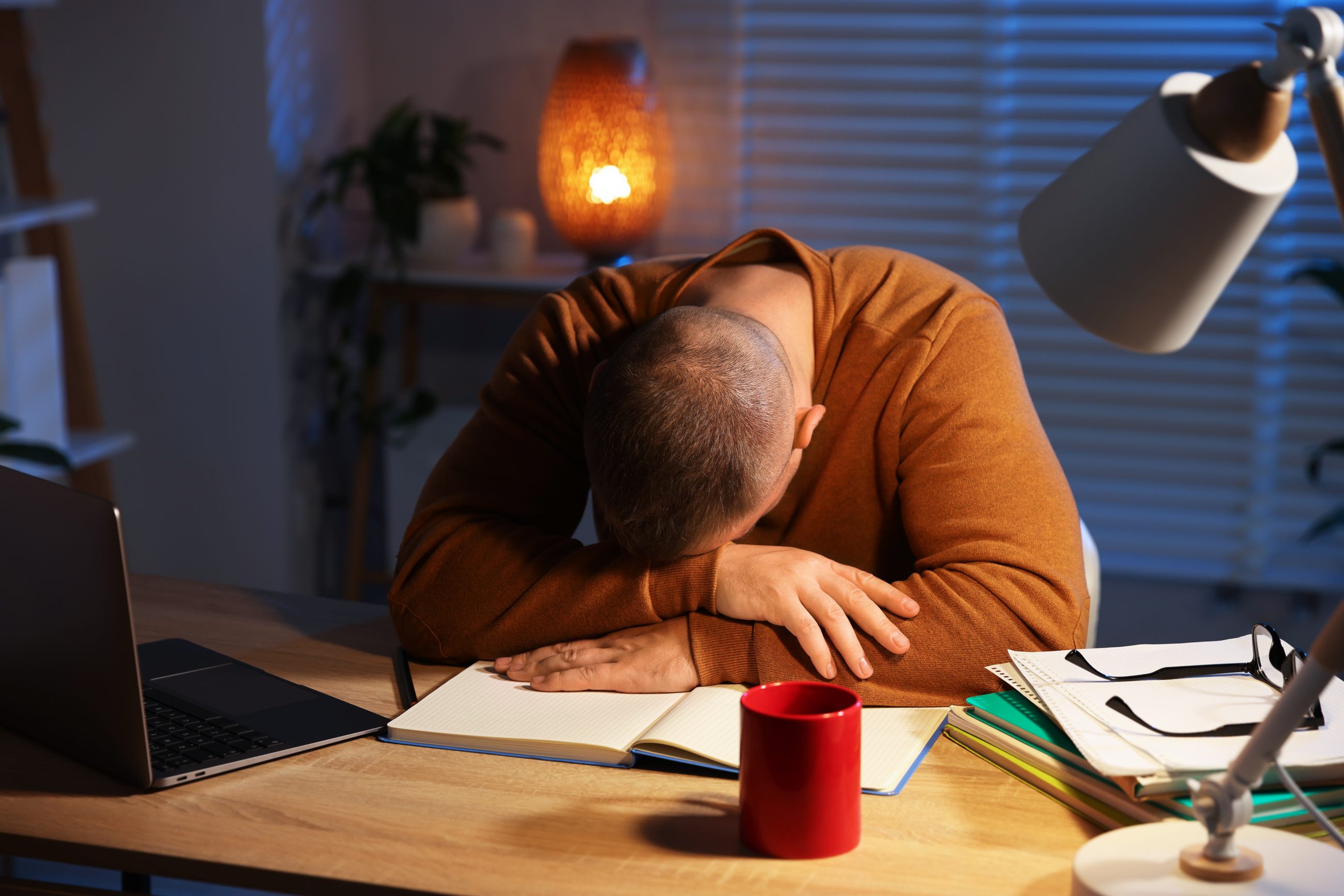 Erschöpfter Mann sitzt spät nachts am Schreibtisch, Kopf auf den Armen abgelegt, als Symbolbild für Functional Freeze