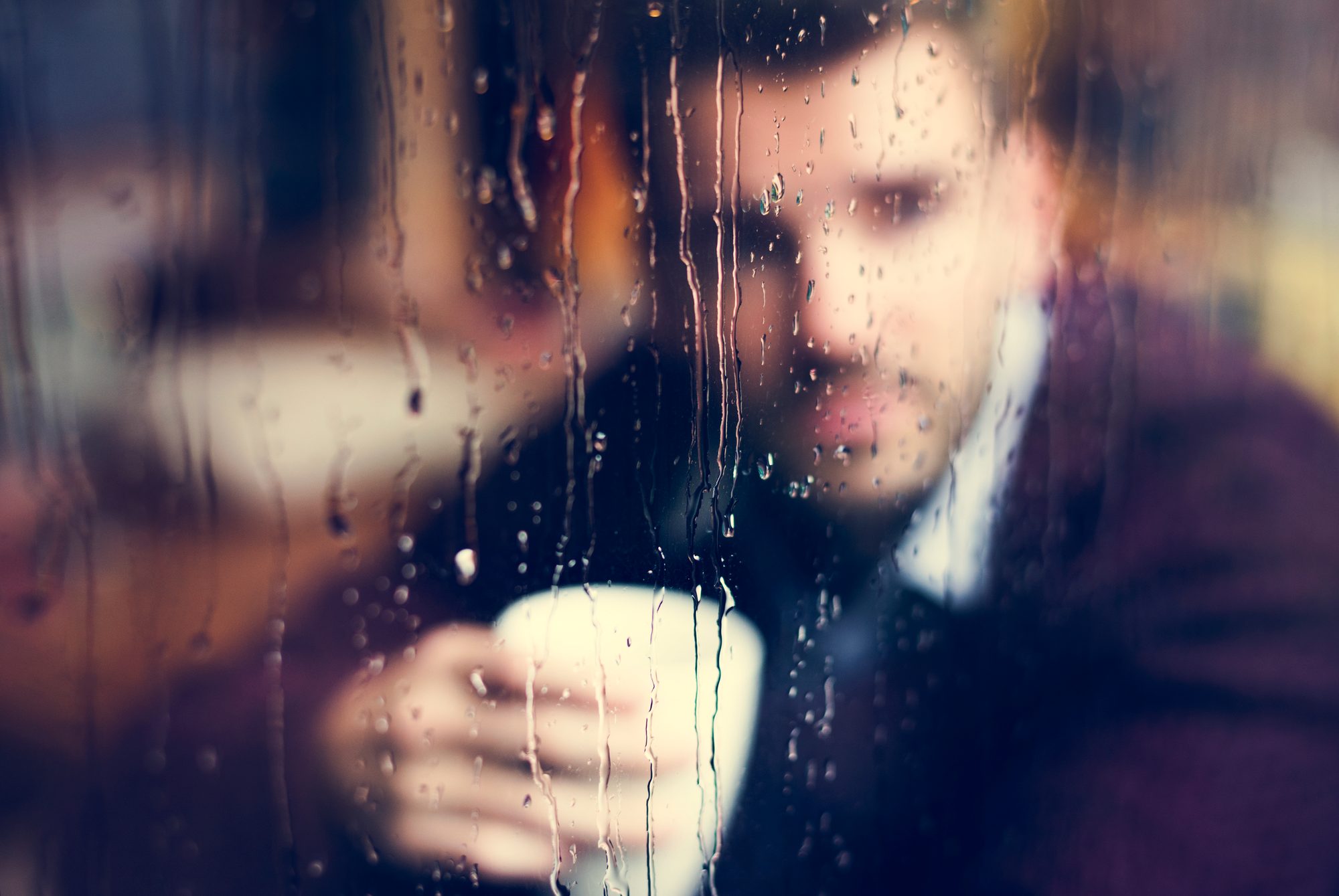 Nachdenklicher Mann mit Tasse hinter einer regennassen Fensterscheibe, Sinnbild für das Grübeln und Warten, das Limerenz kennzeichnet