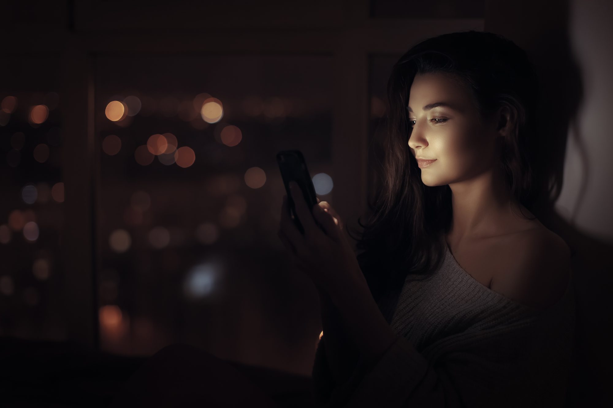 Junge Frau starrt nachts im Halbdunkel auf ihr Smartphone und wartet auf eine Nachricht, Sinnbild für Limerenz und obsessive Verliebtheit