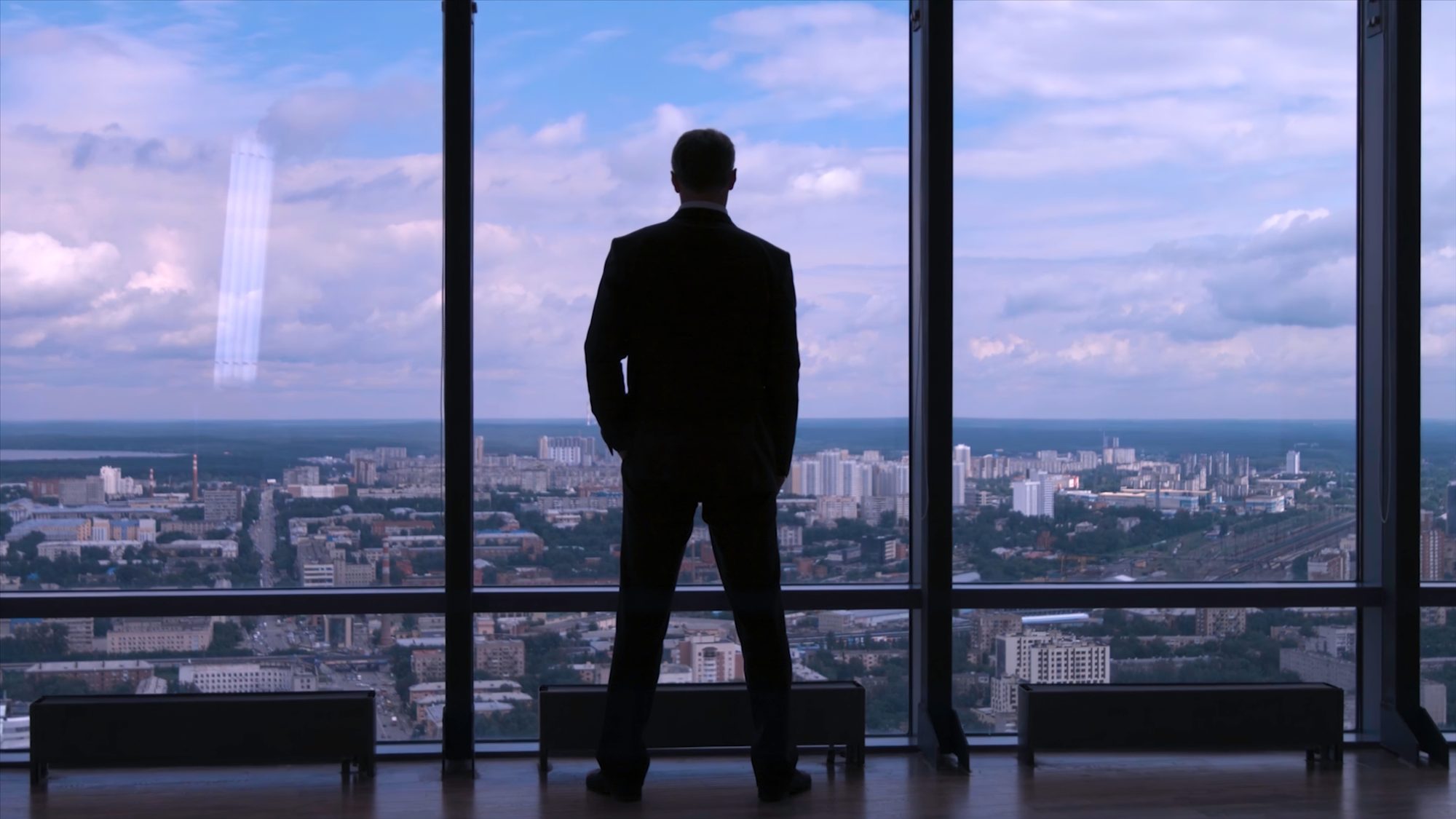 Silhouette eines Managers, der aus dem Bürofenster eines Hochhauses auf die Skyline blickt, Sinnbild für das wandernde Ziel auf der hedonistischen Tretmühle