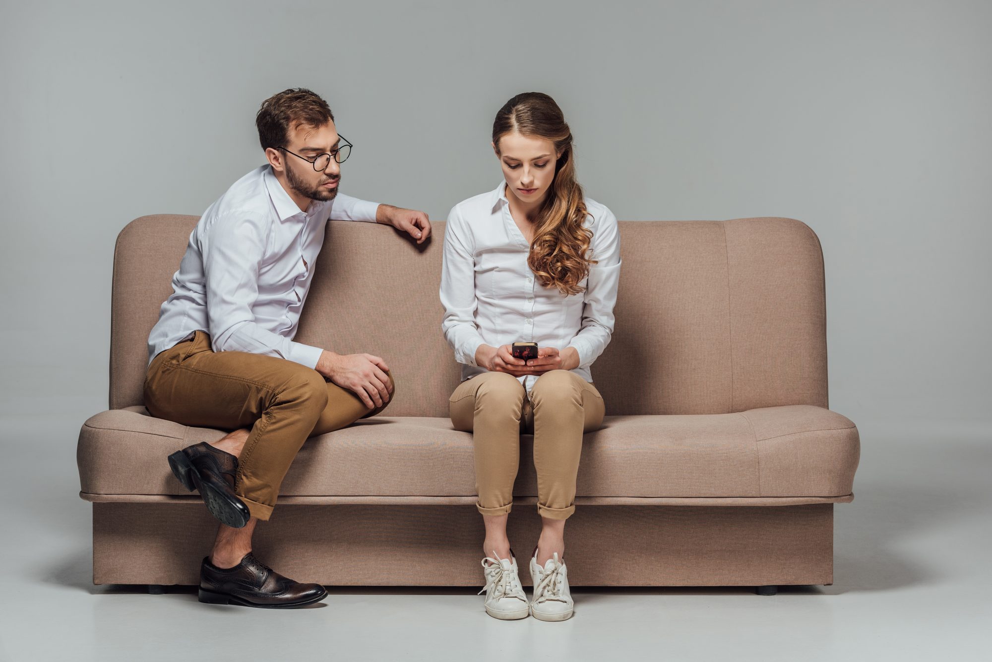 Paar sitzt mit Abstand auf einem Sofa, sie schaut aufs Handy, er blickt sie schweigend an, Sinnbild dafür, die Stille beim Silent Treatment nicht zu füllen