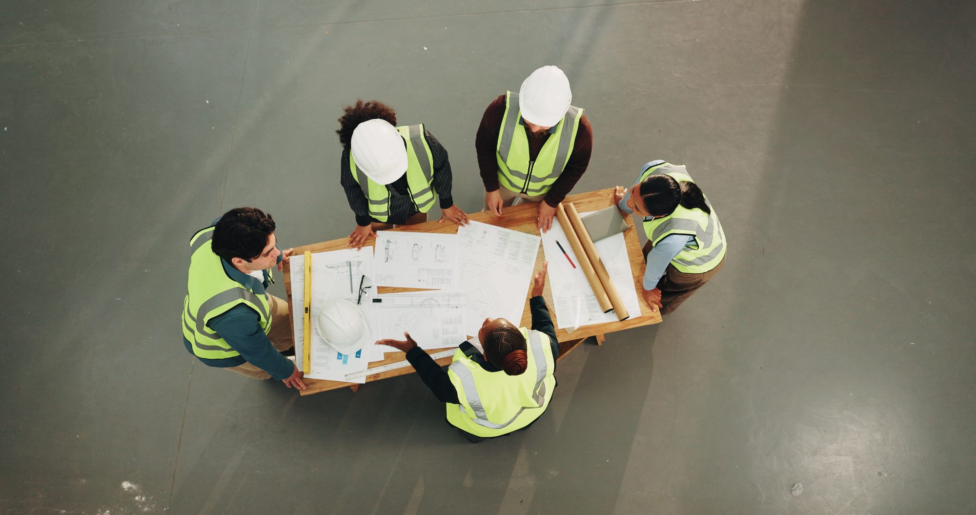 Großes Projektteam beugt sich von oben gesehen über Baupläne auf einem Tisch, Sinnbild für Brooks' Gesetz, nach dem mehr Leute ein verspätetes Projekt weiter verzögern
