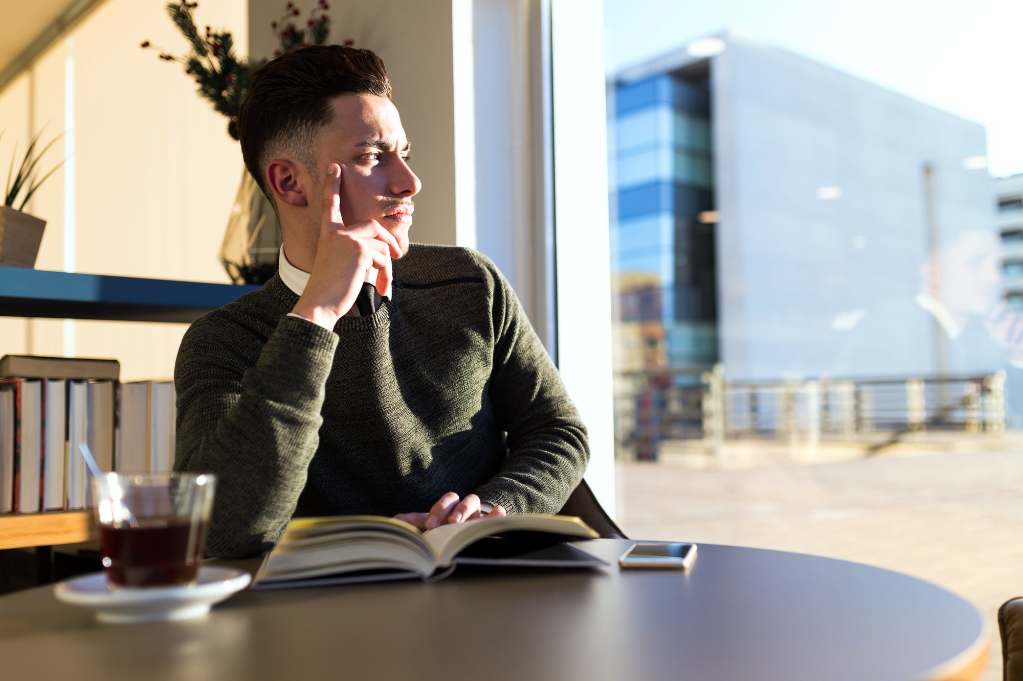 Junger Mann sitzt mit aufgeschlagenem Buch und der Hand am Kinn am Caféfenster, Sinnbild für den Test zur emotionalen Reife