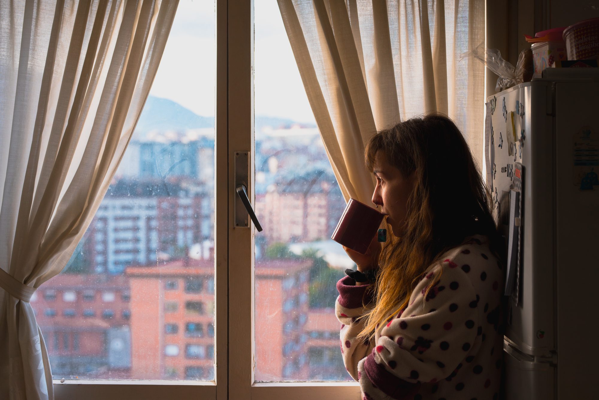 Frau sitzt abends allein mit einer Tasse am Fenster und blickt auf die Stadt, Sinnbild für fehlende Objektkonstanz, wenn der Partner abwesend ist