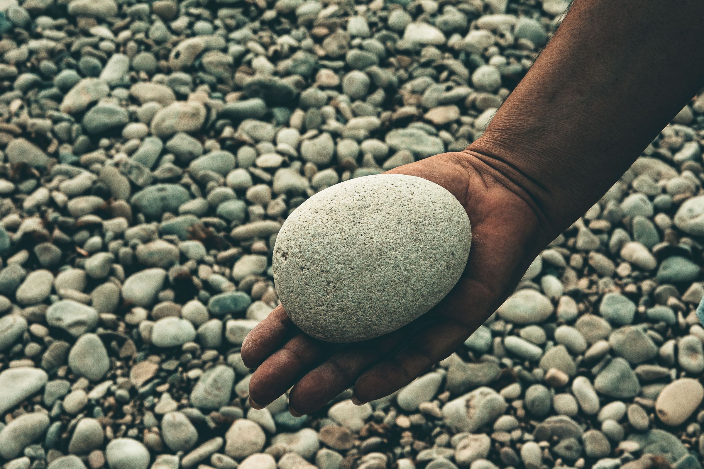 Hand hält einen glatten grauen Kieselstein über einem Kiesstrand, Sinnbild für die Grey Rock Methode im Umgang mit provozierenden Menschen