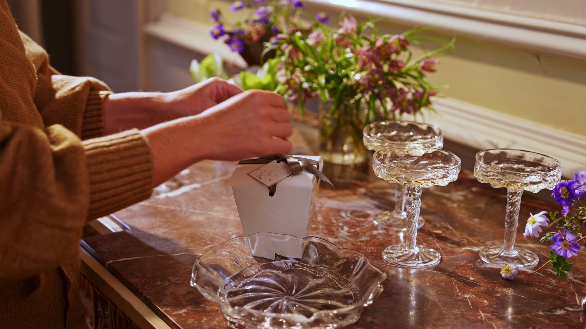 Hænder løsner silkebåndet på en hvid Summerbird-æske på en marmorkamin, omgivet af forårsblomster og champagneglas