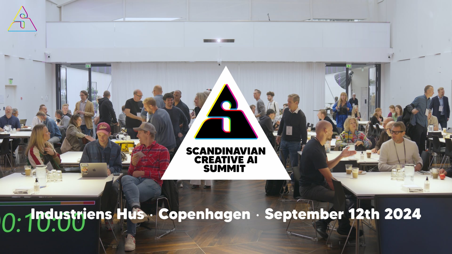 Fyldt konferencesal i Industriens Hus med Scandinavian Creative AI Summit-logoet som en trekant midt i billedet