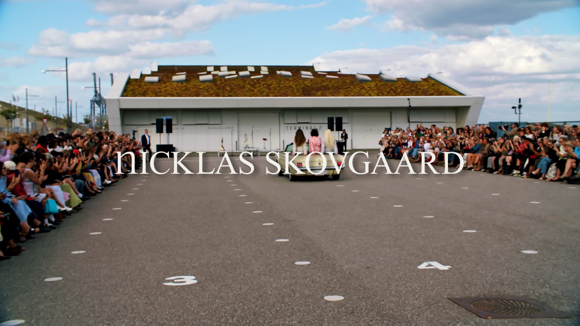 Åbningsbillede fra Nicklas Skovgaards SS27-show foran Terminal 2 i Nordhavn med publikum langs løberen og en vintagebil på vej ind