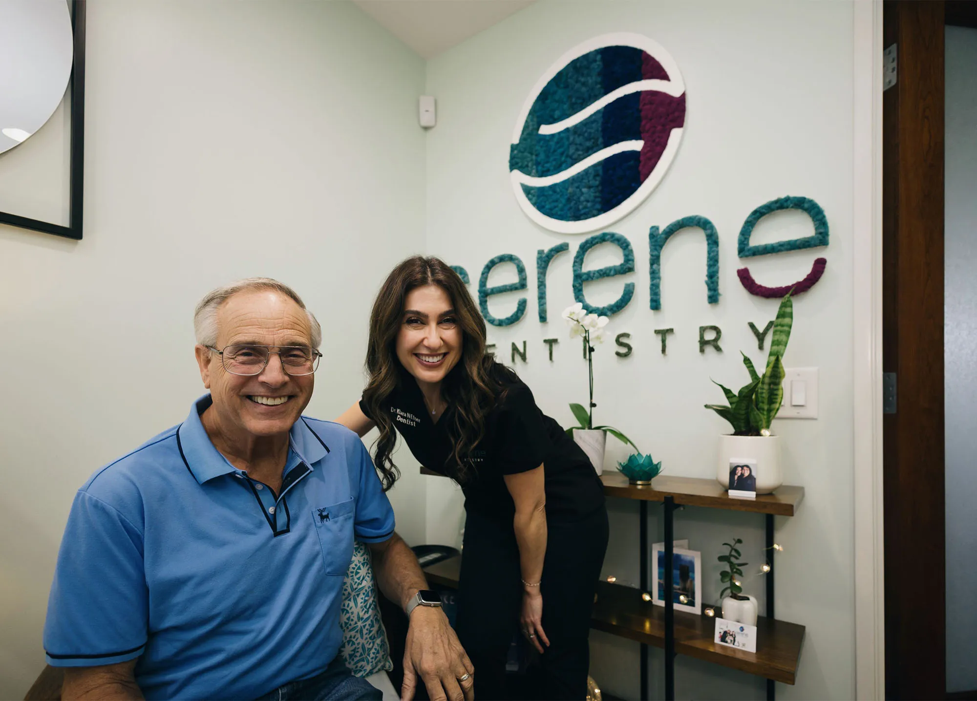 A smiling patient with a Serene Dentistry team member by the practice logo in Ventura, CA