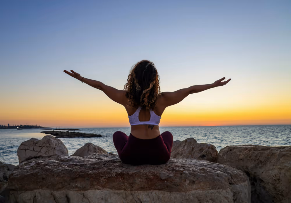 Woman with curly hair sitting cross-legged on a large rock by the sea, arms outstretched at sunset.