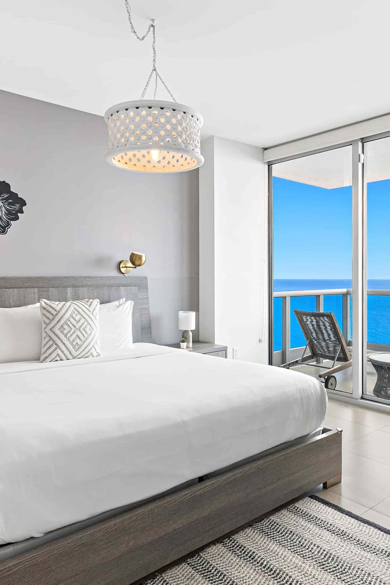 Bedroom at Dharma Home Suites Monte Carlo in Miami Beach with pendant light, gray upholstered headboard, and sliding glass doors opening to a private balcony