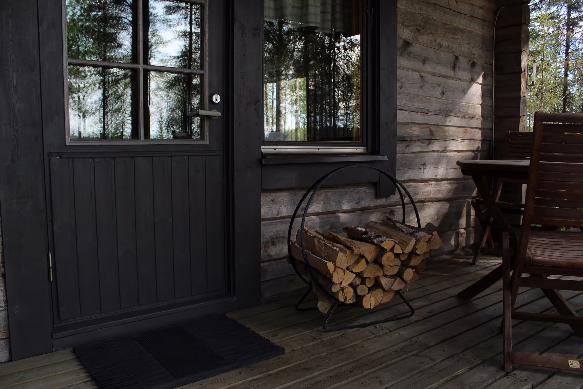 A cozy wooden cabin entrance with a dark door, firewood holder, and an outdoor dining set against a backdrop of tall trees.