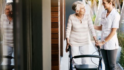 Une femme âgée utilise un déambulateur et elle est aidée par une soignante ou une aide-soignante.