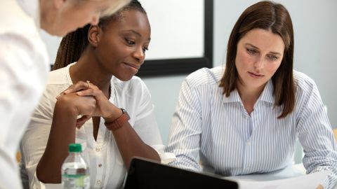 L'image montre trois femmes en train de discuter ensemble autour d'une table dans un cadre de travail ou de réunion.