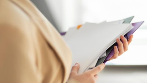 Une personne en tenue professionnelle tient plusieurs dossiers et documents dans les mains.