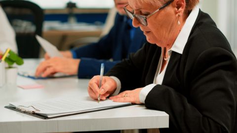 Une femme d'une soixante d'années signe un document posé sur un bureau.
