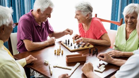 Des personnes âgées souriantes jouent ensemble aux échecs et aux dominos autour d’une table.