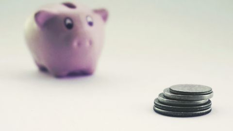 Une tirelire rose floue en arrière-plan et une pile de pièces au premier plan symbolisent l’épargne.
