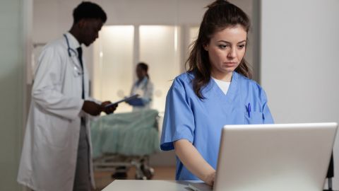 Cadre hospitalier où l'on peut voir trois personnes. Au premier plan, il y a une femme vêtue d'une blouse bleue d'infirmière, concentrée sur l'écran d'un ordinateur portable.