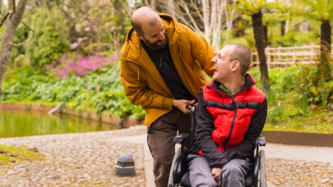 Deux hommes sont dans un parc ou un jardin.L'homme en fauteuil roulant sourit largement en regardant vers le haut vers l'autre homme qui est debout à l’arrière du fauteuil roulant.