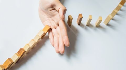 Une main stoppe la chute de dominos en bois, symbolisant la prévention ou la maîtrise d’un risque.