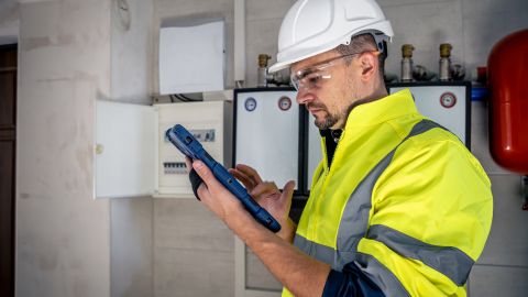 Un homme portant un casque de sécurité blanc et un gilet de sécurité jaune haute visibilité effectuant une inspection. Il regarde une tablette qu'il tient dans ses mains.