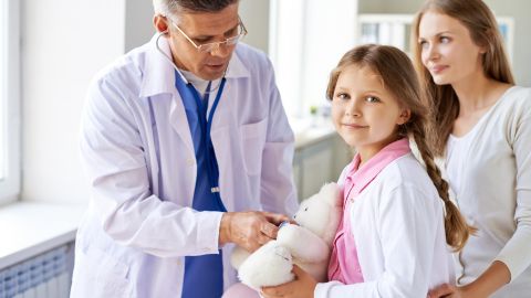 Un médecin ausculte un ours en peluche tenu par une fillette, sous le regard bienveillant de sa mère.
