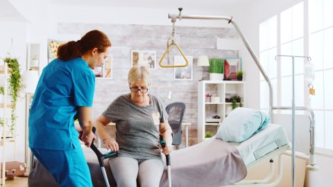 Sur cette image, on voit une infirmière aider une patiente âgée à se lever de son lit médicalisé en soins à domicile.