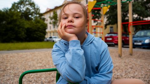Petite fille atteinte de trisomie 21 assise sur un équipement de jeu dans un parc. Elle semble songeuse, appuyant sa tête sur sa main gauche avec un regard sérieux ou pensif.