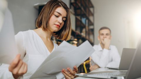 Une femme en chemise blanche examine des documents dans un bureau, tandis qu'un homme assis derrière elle semble réfléchir.