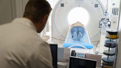 Patient en train de passer un examen dans un appareil de type IRM ou un scanner. Le patient est allongé sur une table mobile qui est partiellement entrée dans le large anneau de l'appareil. En premier plan, un professionnel de santé, observe l'examen depuis une salle de contrôle, où plusieurs écrans et équipements de surveillance sont visibles.