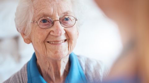 Dame âgée souriante portant des lunettes avec des montures fines aux cheveux blancs coiffés soigneusement. Elle fait face à une femme qui est en flou au premier plan.