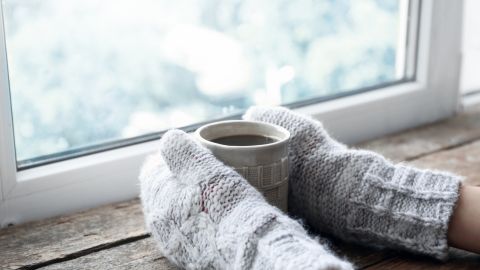 L'image montre des mains couvertes de gants gris en tricot épais, tenant une tasse de boisson chaude près d'une fenêtre.